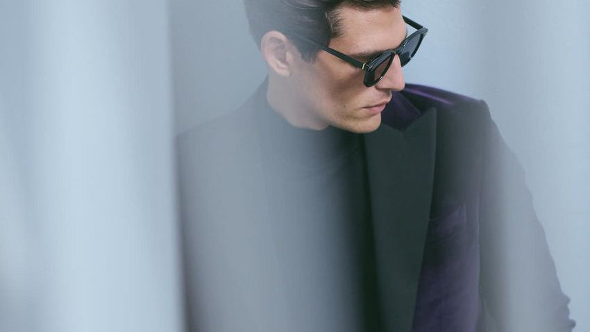 A man wearing black sunglasses, a black turtleneck, and a dark purple blazer draped over his shoulder, posed against a soft, light gray background with a blurred foreground overlay. Sand Copenhagen Suits available at Sakks Menswear in Southampton.