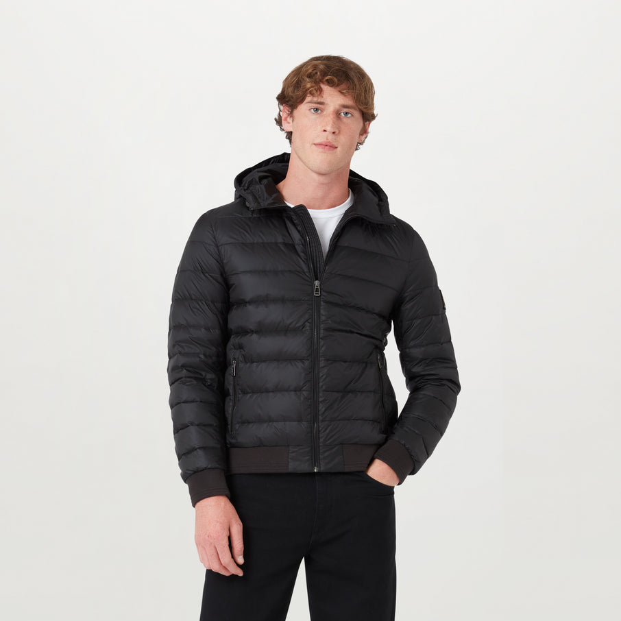 A man wearing a black hooded puffer jacket with a full-zip front, ribbed cuffs and hem, and a white t-shirt underneath, standing against a light grey studio background. Belstaff Down Jacket. Sakks Menswear Southampton.