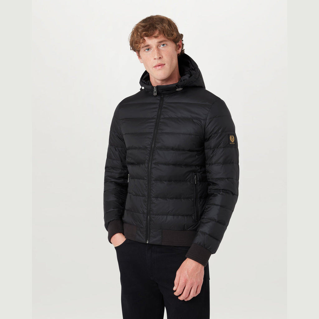 A man wearing a black hooded puffer jacket with a full-zip front, ribbed cuffs and hem, and a small yellow embroidered logo patch on the left sleeve, photographed against a plain light grey studio background. Belstaff Down Jacket. Sakks Menswear Southampton.