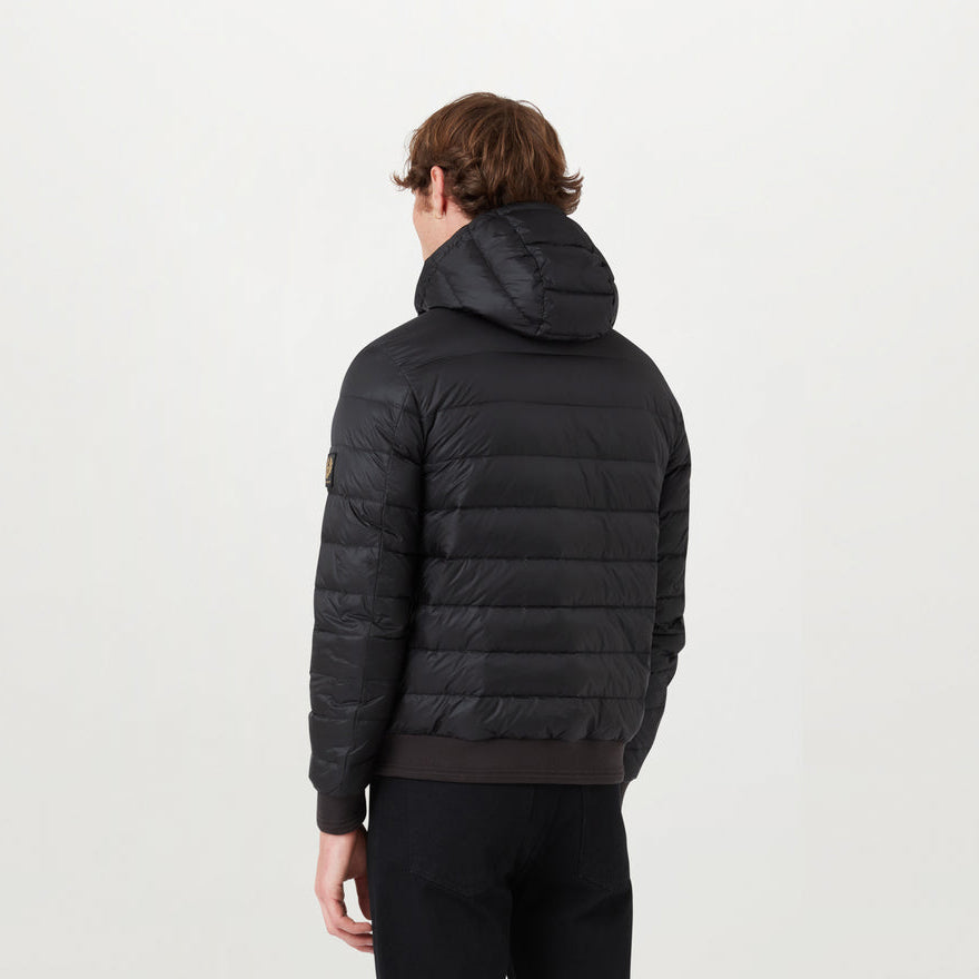 A man viewed from the back wearing a black hooded puffer jacket with horizontal quilting and a small tan logo patch on the upper left sleeve, set against a plain white studio background. Belstaff Down Jacket. Sakks Menswear Southampton.