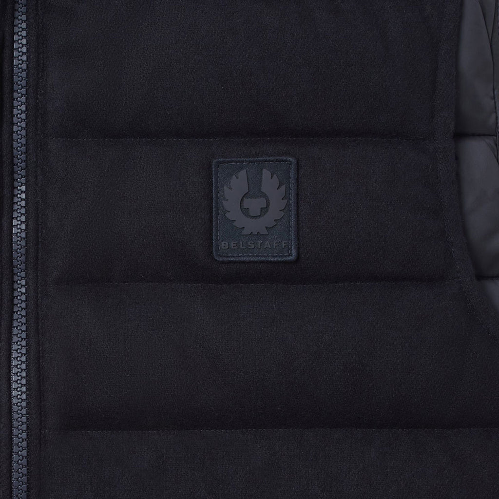 Close-up of a black puffer vest showing the central zip closure and a dark gray rectangular Beaufort brand patch on the chest. Belstaff Gilet. Sakks Menswear Southampton.