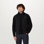 A man wearing a black quilted puffer vest over a black ribbed knit sweater and dark blue jeans, standing against a plain light gray studio background. Belstaff Gilet. Sakks Menswear Southampton.