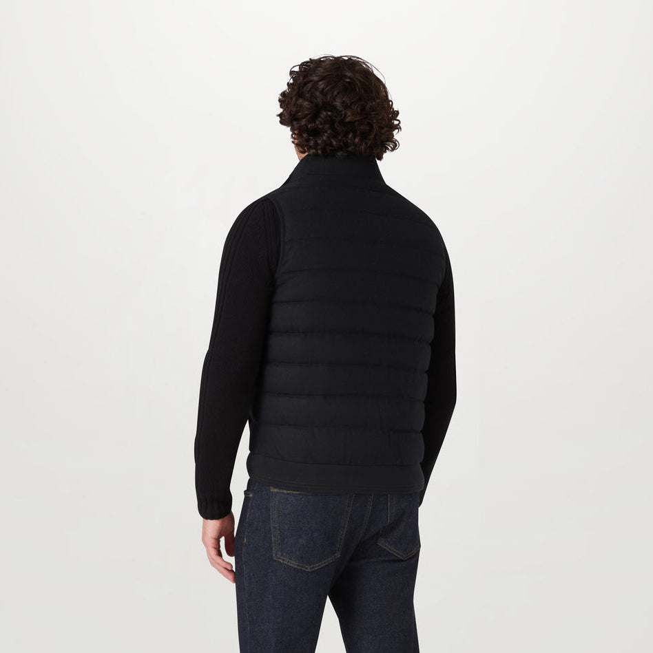 A man viewed from the back wearing a black quilted puffer vest over a black long-sleeve top and dark blue jeans, against a plain light gray studio background. Belstaff Gilet. Sakks Menswear Southampton.