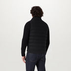 A man viewed from the back wearing a black quilted puffer vest over a black long-sleeve top and dark blue jeans, against a plain light gray studio background. Belstaff Gilet. Sakks Menswear Southampton.