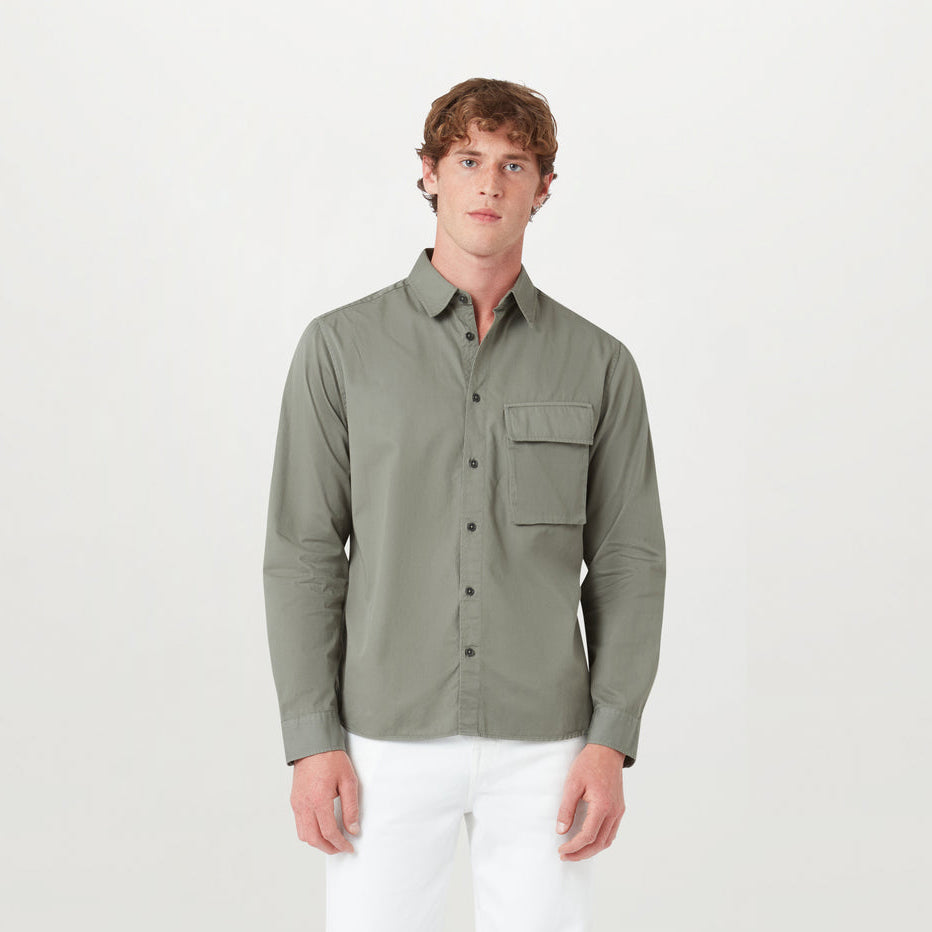 A man wearing a long-sleeve button-up shirt in sage green with a chest pocket, paired with white pants, standing against a plain light gray background. Belstaff Shirt. Sakks Menswear Southampton.