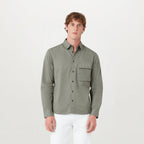 A man wearing a long-sleeve button-up shirt in sage green with a chest pocket, paired with white pants, standing against a plain light gray background. Belstaff Shirt. Sakks Menswear Southampton.