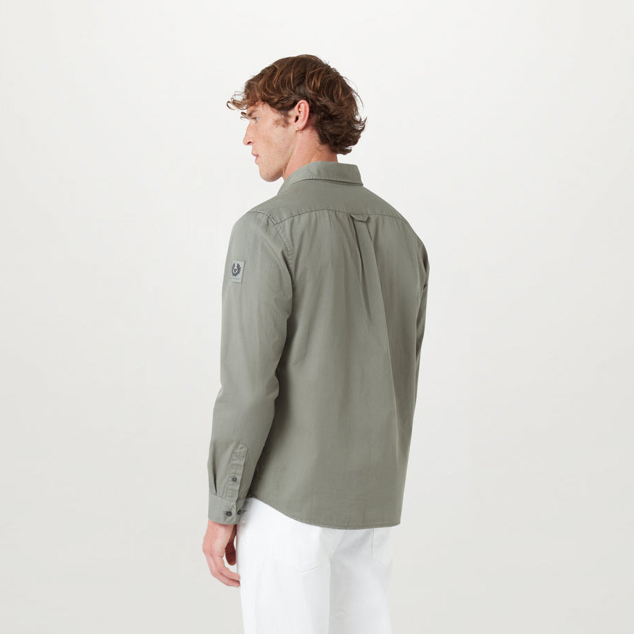 A man wearing a sage green long-sleeve button-up shirt and white trousers, shown from a side-back angle against a plain light gray studio background. Belstaff Shirt. Sakks Menswear Southampton.