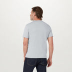 A man viewed from behind wearing a light gray short-sleeve t-shirt and dark blue jeans, standing against a plain white studio background. Belstaff T-Shirt. Sakks Menswear Southampton.