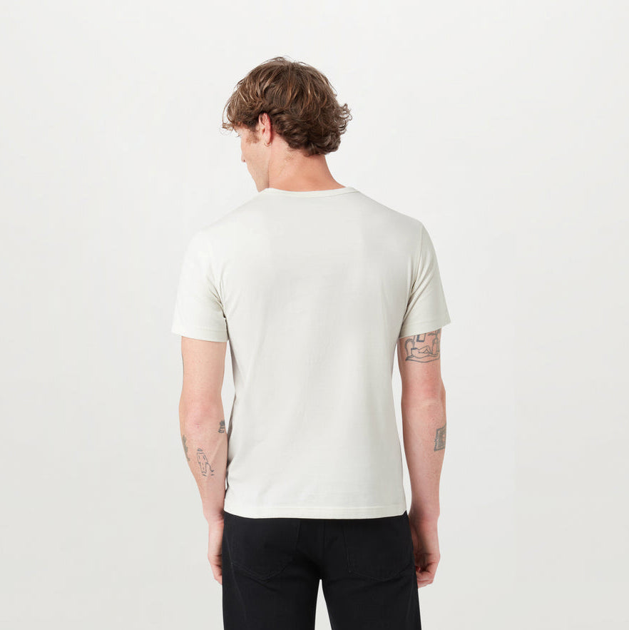 A man viewed from behind wearing a light cream/off-white short-sleeve crewneck t-shirt and black pants, standing against a plain white studio background. Belstaff T-Shirt. Sakks Menswear Southampton.