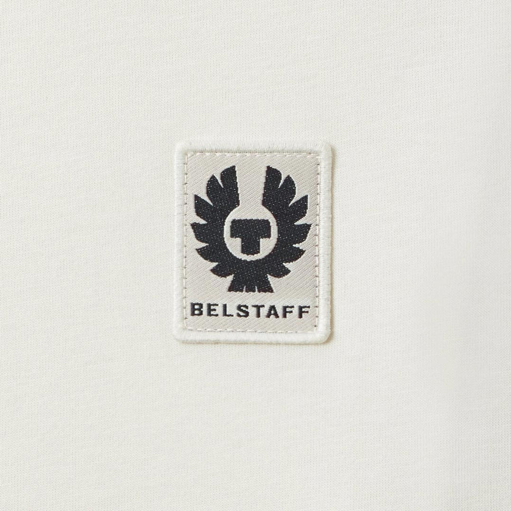 A close-up of a white rectangular embroidered patch featuring the black Belstaff winged logo and the brand name 'BELSTAFF' on a white textured fabric background. Belstaff T-Shirt. Sakks Menswear Southampton.