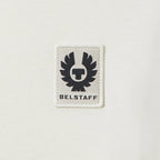 A close-up of a white rectangular embroidered patch featuring the black Belstaff winged logo and the brand name 'BELSTAFF' on a white textured fabric background. Belstaff T-Shirt. Sakks Menswear Southampton.