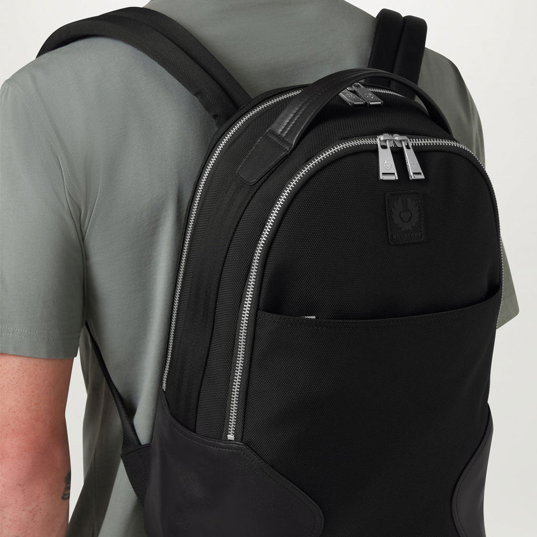 A black backpack with silver zippers worn by a person wearing a gray t-shirt, shown from a rear three-quarter angle against a light gray background. Belstaff Backpack. Sakks Menswear Southampton