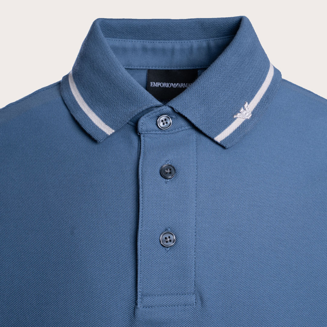 Close-up of a blue polo shirt collar with white stripe trim, three dark buttons, and a small white embroidered logo on the collar, shown against a light neutral background. Emporio Armani Polo. Sakks Menswear Southampton.