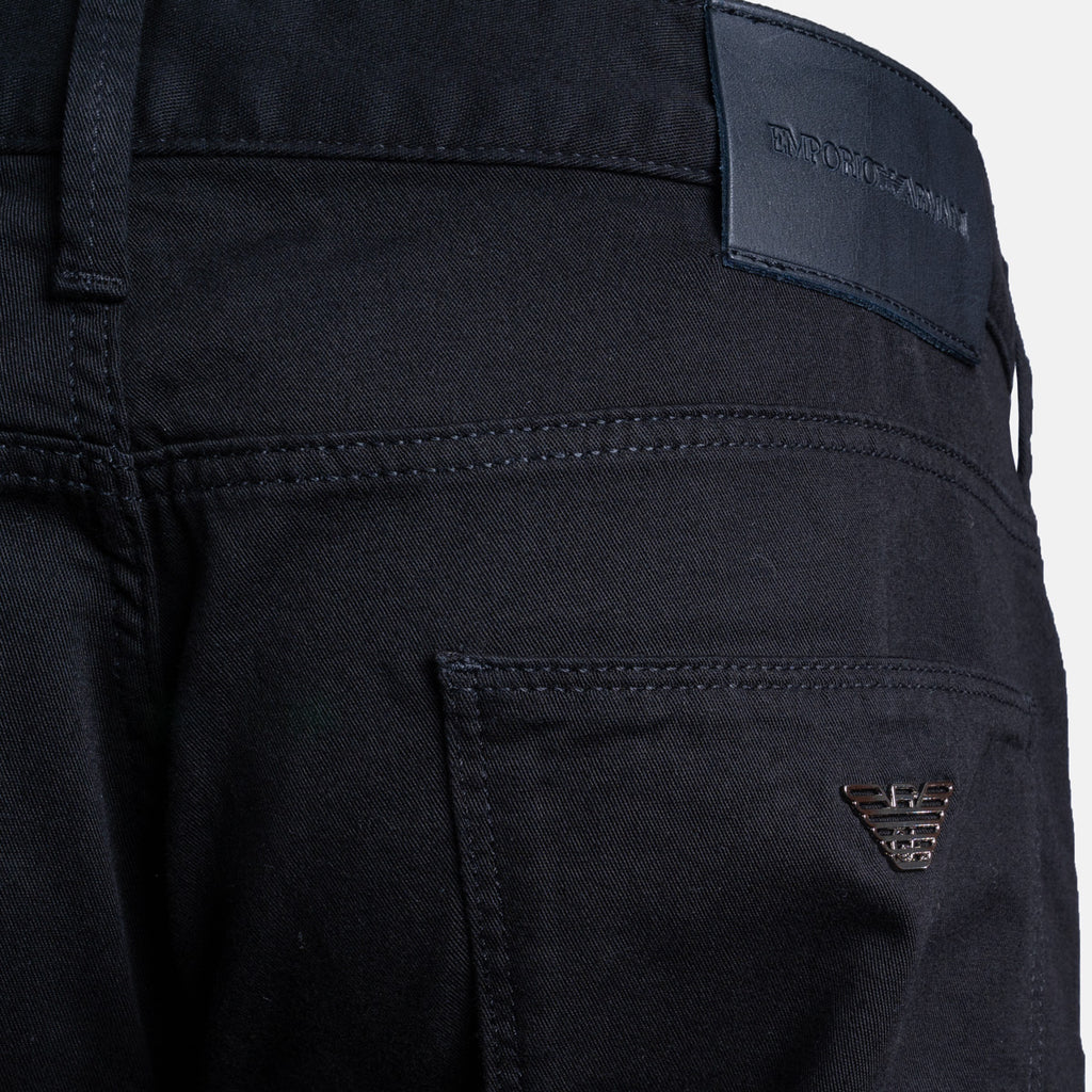 Close-up of the back waistband and pocket area of dark navy blue denim jeans, showing a black leather brand patch with debossed text and a small metallic Emporio Armani eagle logo on the pocket. Emporio Armani Chinos. Sakks Menswear Southampton.
