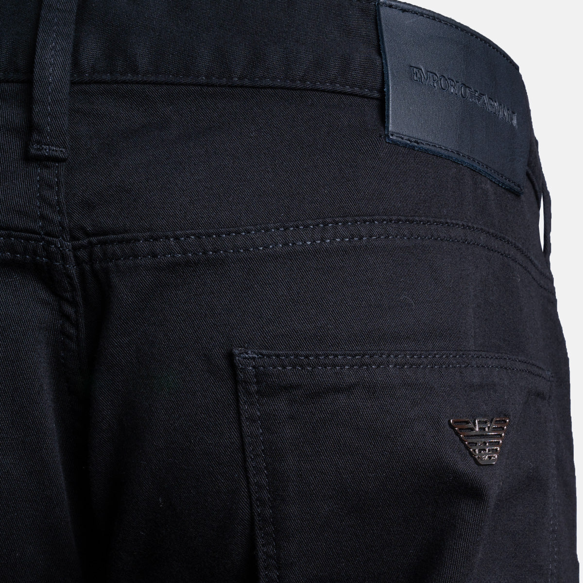 Close-up of the back waistband and pocket area of dark navy blue denim jeans, showing a black leather brand patch with debossed text and a small metallic Emporio Armani eagle logo on the pocket. Emporio Armani Chinos. Sakks Menswear Southampton.