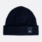 A dark navy blue knit beanie hat with a folded cuff, featuring a small white embroidered Emporio Armani eagle logo patch on the right side of the cuff, set against a light gray background. Emporio Armani Beanie. Sakks Menswear Southampton.