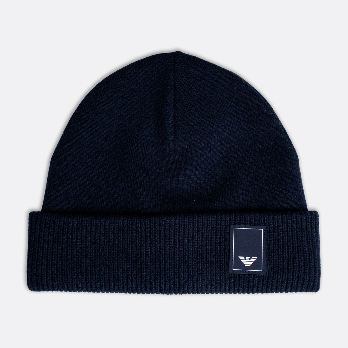 A dark navy blue knit beanie hat with a folded cuff, featuring a small white embroidered Emporio Armani eagle logo patch on the right side of the cuff, set against a light gray background. Emporio Armani Beanie. Sakks Menswear Southampton.