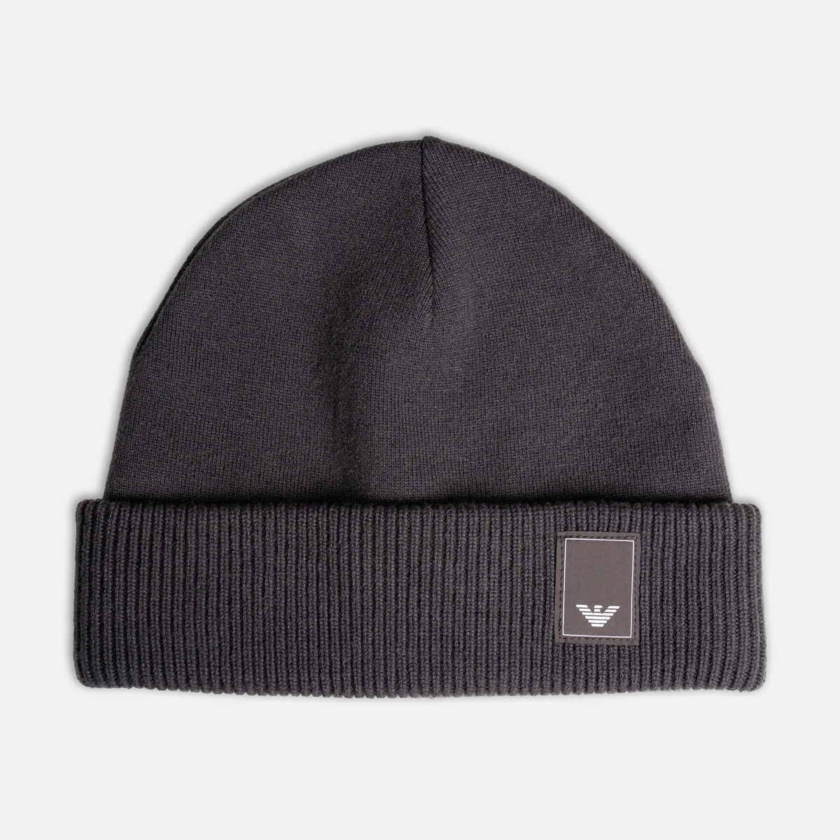 A dark gray ribbed knit beanie hat with a folded cuff and a small square patch featuring the Emporio Armani eagle logo, shown against a light gray background. Emporio Armani Beanie. Sakks Menswear Southampton.