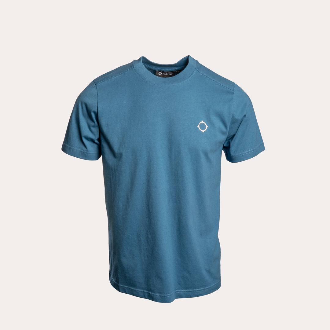 A blue short-sleeve crewneck t-shirt with a small white geometric logo on the left chest, displayed on a light beige background. MA.Strum T-Shirt available at Sakks Menswear in Southampton.