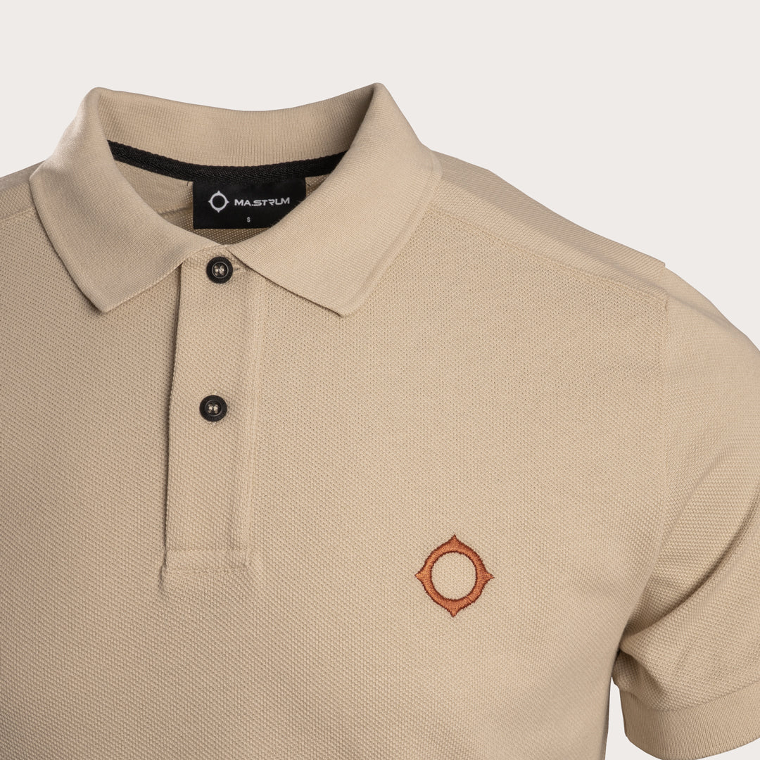 A close-up of a beige short-sleeve polo shirt with a collar, two-button placket, and a small embroidered circular logo on the left chest, shown against a light neutral background. MA.Strum Polo available at Sakks Menswear in Southampton.