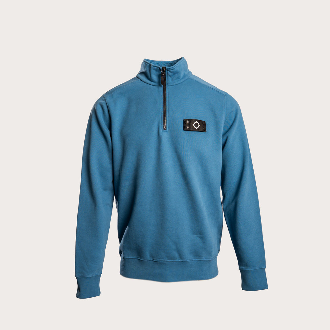 A blue long-sleeve quarter-zip sweatshirt with a mock neck collar and a black rectangular logo patch on the left chest, displayed on a neutral light beige background. MA.Strum Sweatshirt available at Sakks Menswear in Southampton.