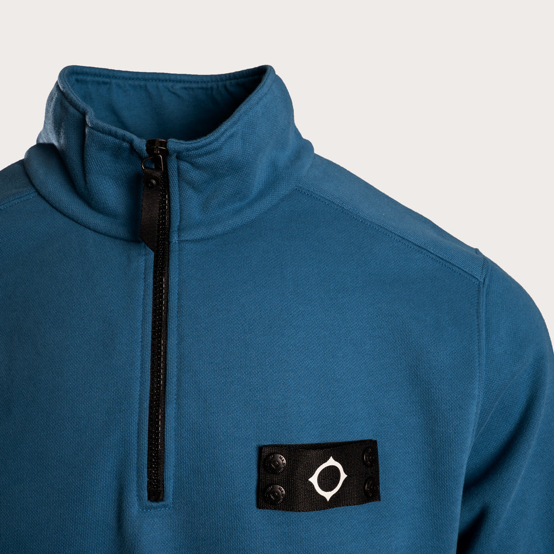 A close-up of a blue quarter-zip fleece pullover with a black zipper, a black buttoned patch on the chest featuring a white compass-like logo, and a light gray background. MA.Strum Sweatshirt available at Sakks Menswear in Southampton.
