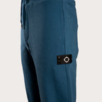Close-up of a pair of blue pants showing a black fabric brand patch with a white compass design on the side pocket. MA.Strum Sweat Pants available at Sakks Menswear in Southampton.