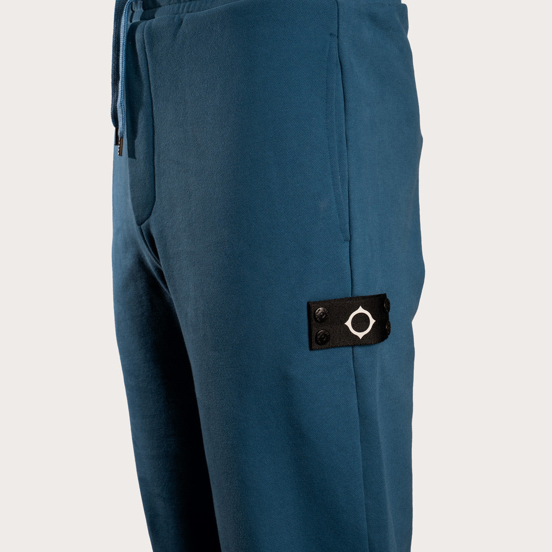 Close-up of a pair of blue pants showing a black fabric brand patch with a white compass design on the side pocket. MA.Strum Sweat Pants available at Sakks Menswear in Southampton.