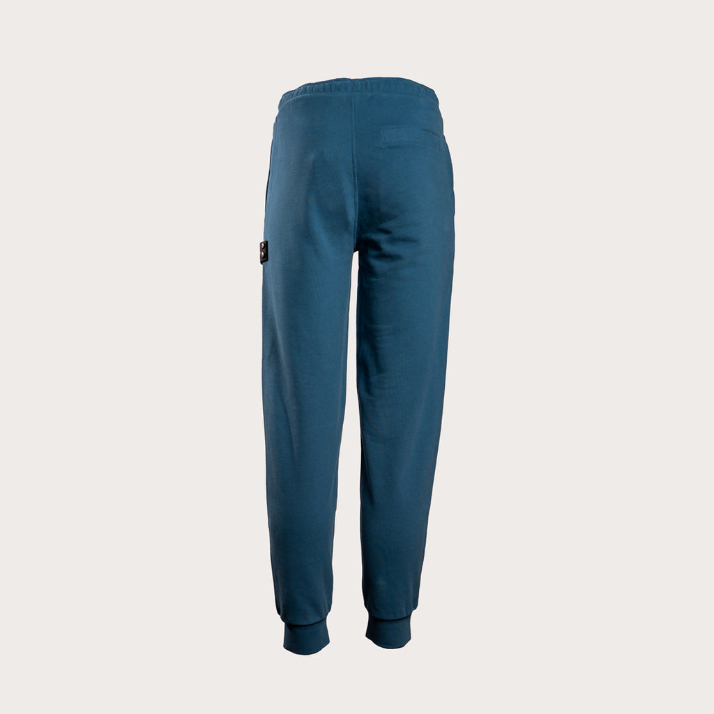 A pair of dark teal blue sweatpants shown from the back, featuring a small black brand patch on the left side, set against a solid light gray background. MA.Strum Sweat Pants available at Sakks Menswear in Southampton.