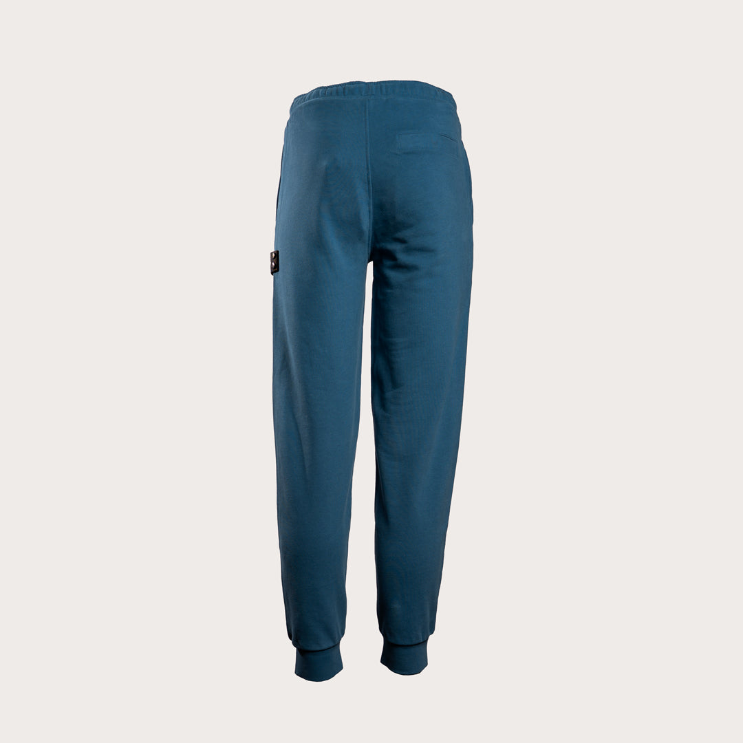 A pair of dark teal blue sweatpants shown from the back, featuring a small black brand patch on the left side, set against a solid light gray background. MA.Strum Sweat Pants available at Sakks Menswear in Southampton.