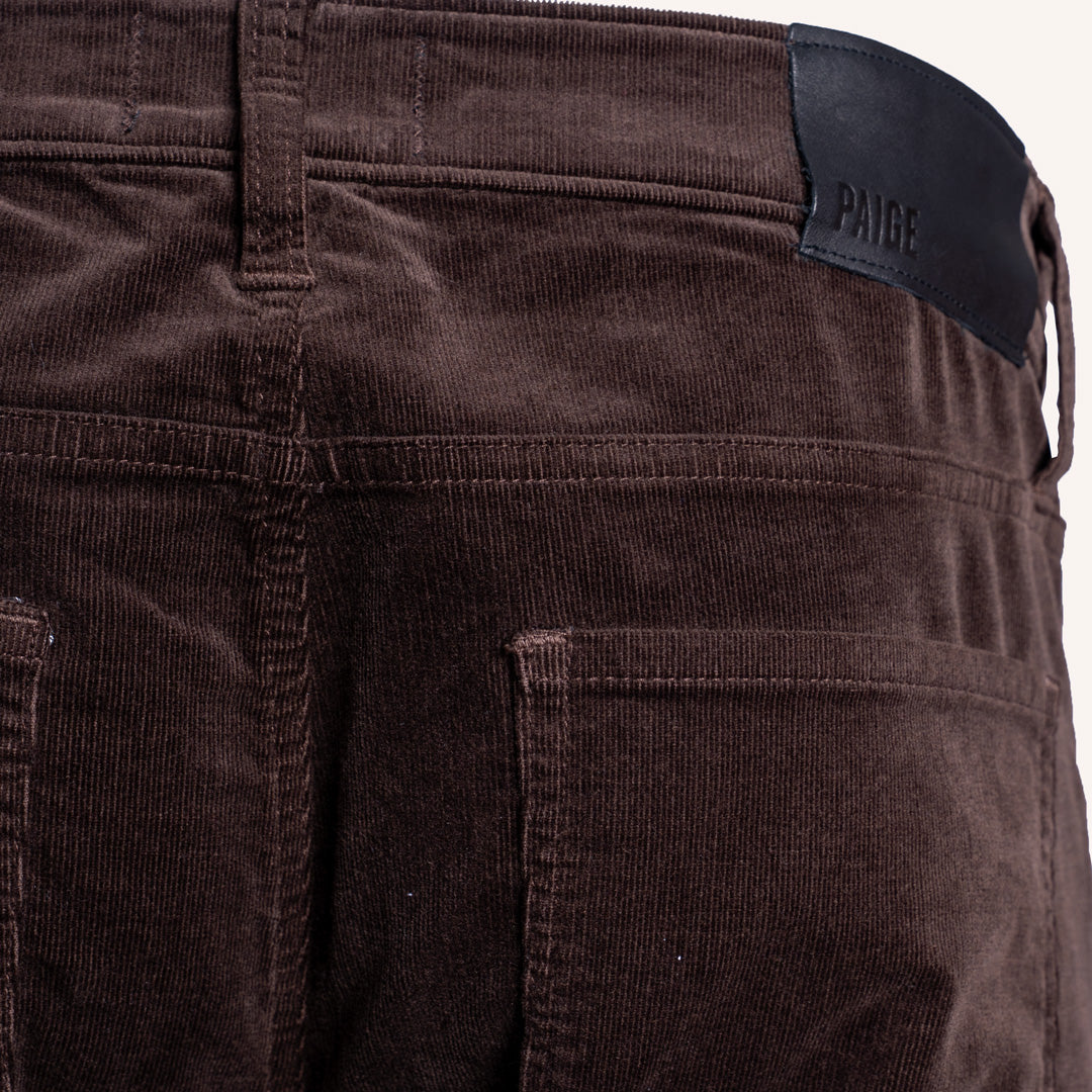 Close-up of the back waistband of dark brown corduroy pants, showing a black leather brand patch with the embossed brand name PAIGE. PAIGE Jeans available at Sakks Menswear in Southampton.