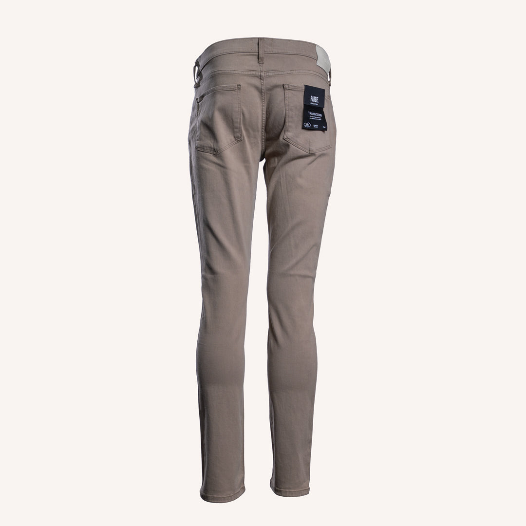 A pair of slim-fit jeans in a taupe-brown color, shown from the back on a light cream background. A black brand label is visible on the back right pocket. PAIGE Jeans available at Sakks Menswear in Southampton.