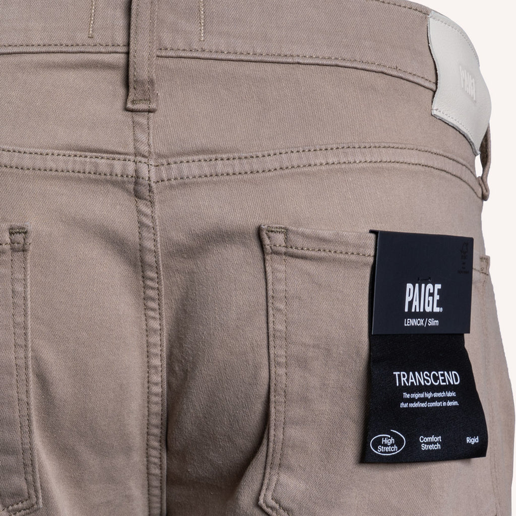 Close-up of the back pocket area of Paige Lexington 510m denim jeans in a muted taupe-brown color, featuring a black brand label on the pocket. PAIGE Jeans available at Sakks Menswear in Southampton.