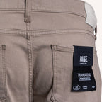 Close-up of the back pocket area of Paige Lexington 510m denim jeans in a muted taupe-brown color, featuring a black brand label on the pocket. PAIGE Jeans available at Sakks Menswear in Southampton.