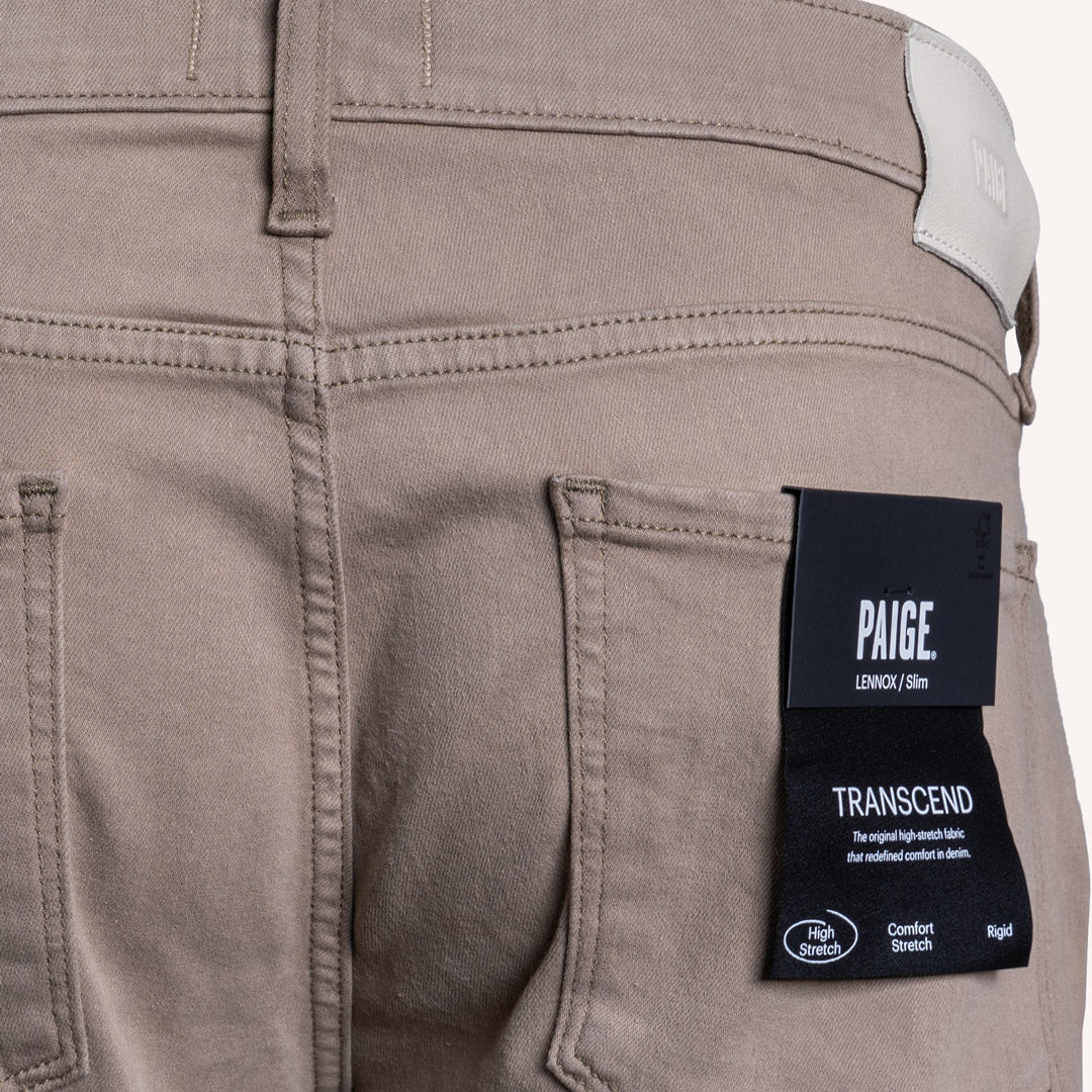Close-up of the back pocket area of Paige Lexington 510m denim jeans in a muted taupe-brown color, featuring a black brand label on the pocket. PAIGE Jeans available at Sakks Menswear in Southampton.