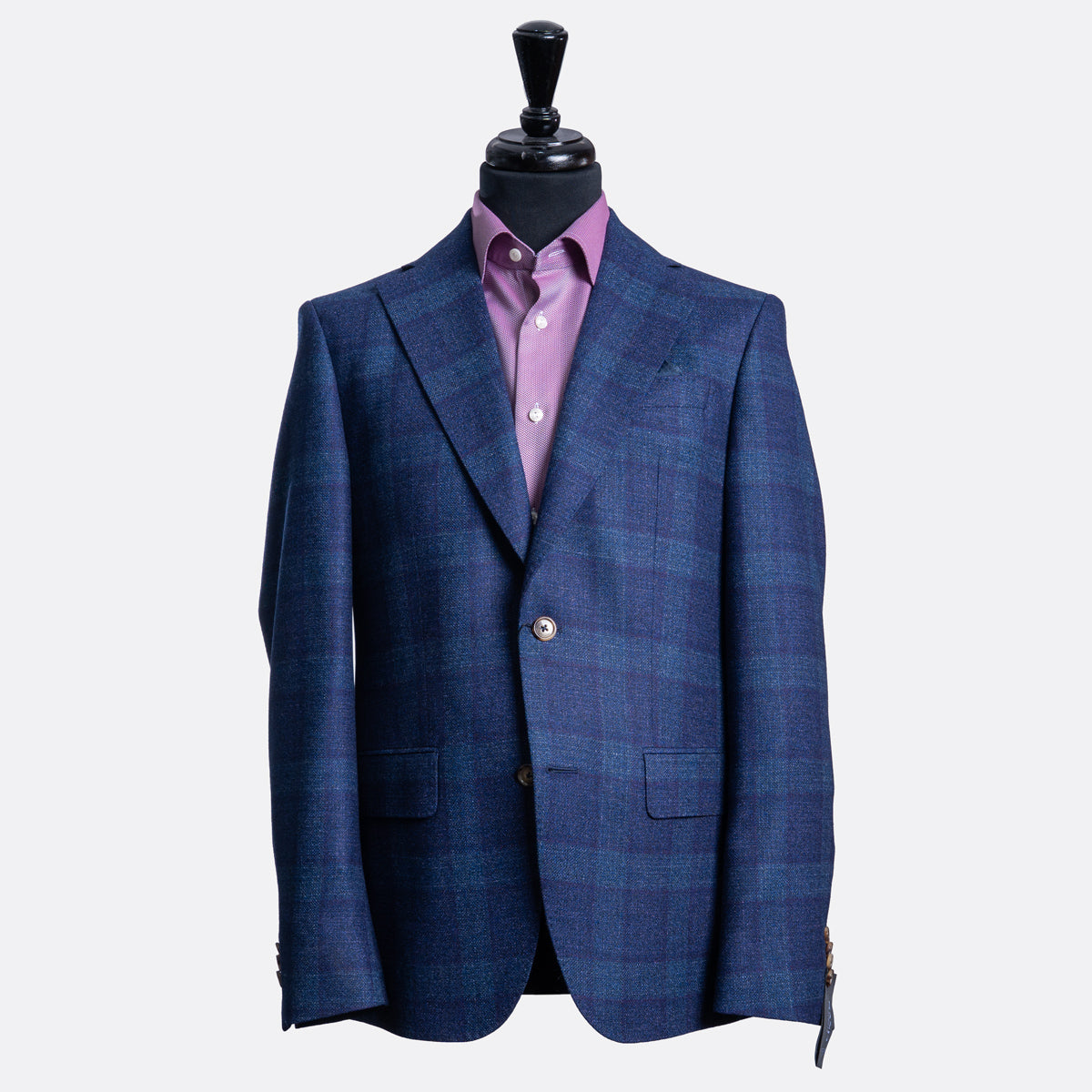 A blue and navy plaid blazer displayed on a black mannequin torso, layered over a purple dress shirt, against a light gray background. Sand Copenhagen Blazer available at Sakks Menswear in Southampton.
