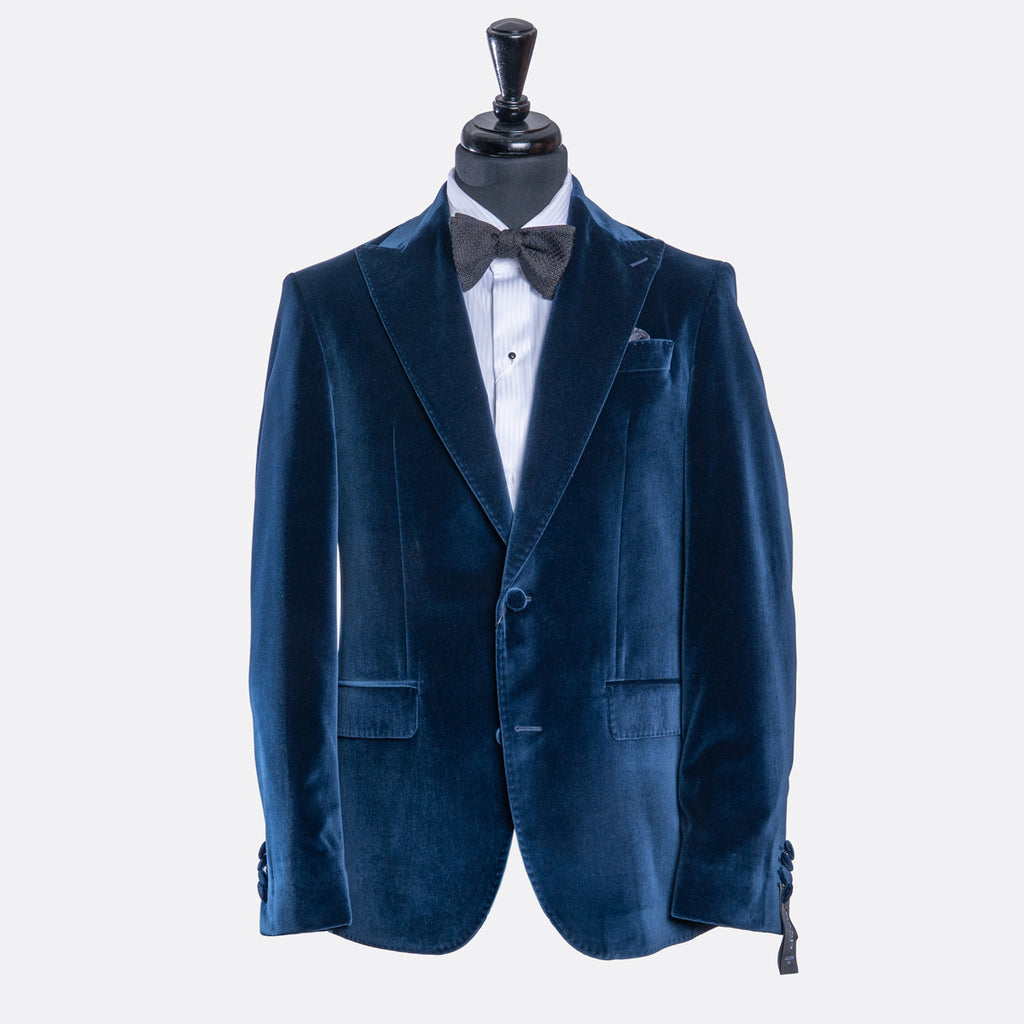 A men's navy blue velvet blazer displayed on a black mannequin torso, styled with a white dress shirt, a dark gray bow tie, and a white pocket square against a light gray background. Sand Copenhagen Velvet Blazer available at Sakks Menswear in Southampton.