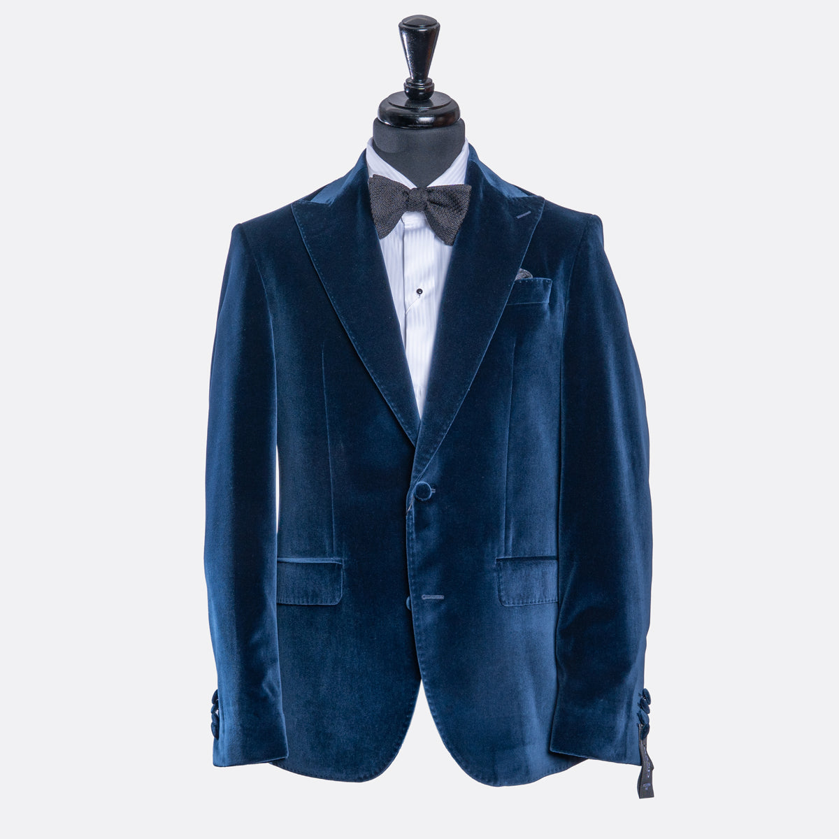 A men's navy blue velvet blazer displayed on a black mannequin torso, styled with a white dress shirt, a dark gray bow tie, and a white pocket square against a light gray background. Sand Copenhagen Velvet Blazer available at Sakks Menswear in Southampton.