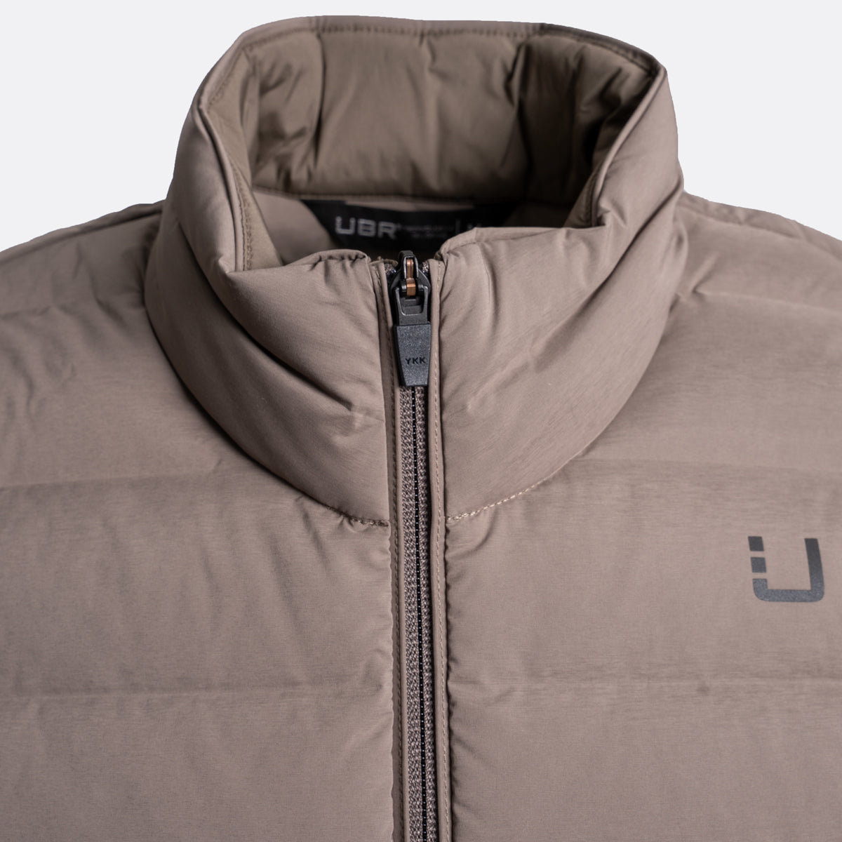 Close-up of a puffer jacket in taupe-brown with a high collar, front zipper, and a dark gray stylized logo on the chest. UBR Gilet available at Sakks Menswear in Southampton.