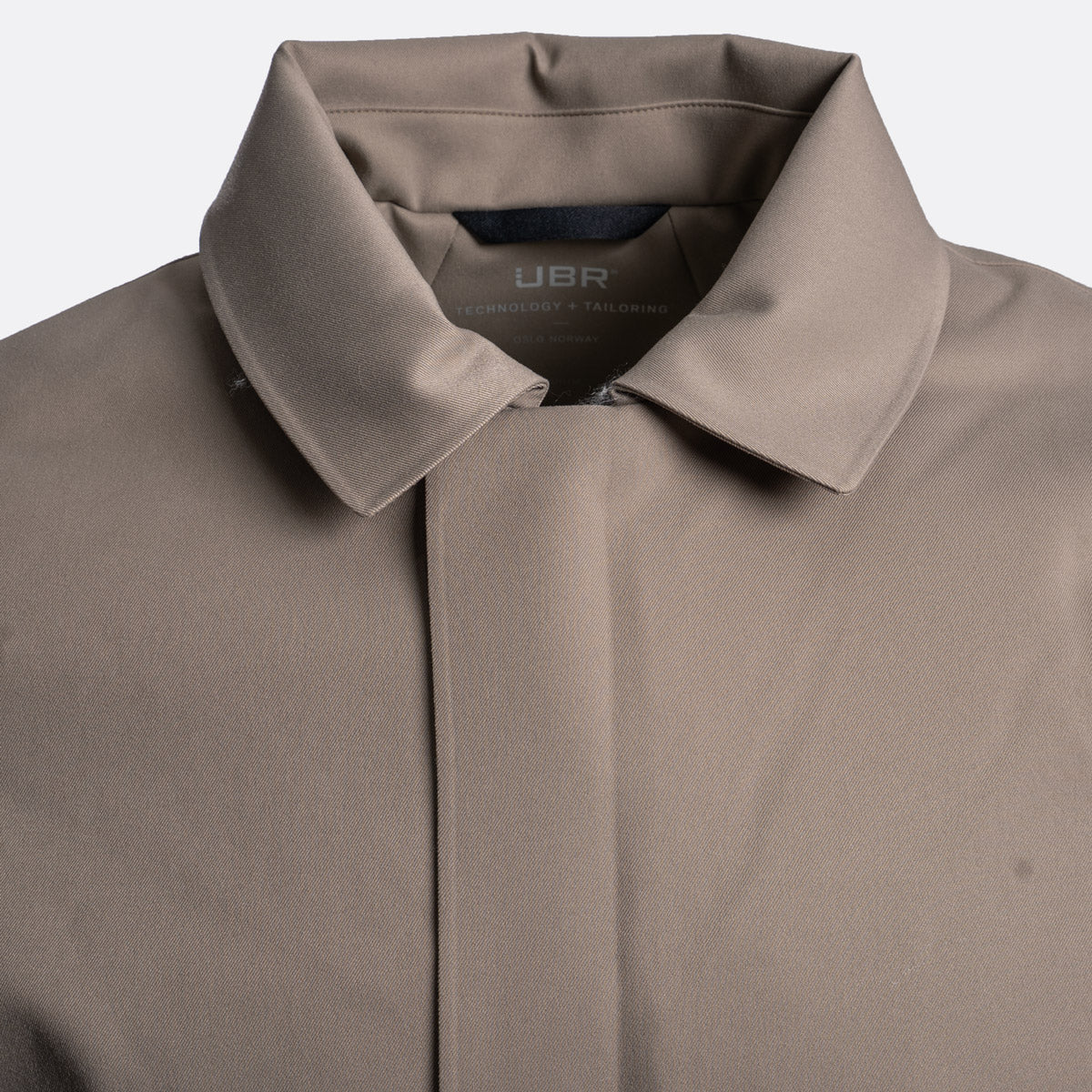 Close-up of a taupe-colored collared shirt by UBR, showing the collar, inner neck label, and front placket against a light gray background. UBR Jacket available at Sakks Menswear in Southampton.
