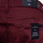 Close-up of the back waistband and pocket area of burgundy slim-fit pants, showing a black fabric brand label reading 'SLIMMY' and a black hangtag reading 'LUXE PERFORMANCE'. 7 For All Mankind. Sakks Menswear Southampton.
