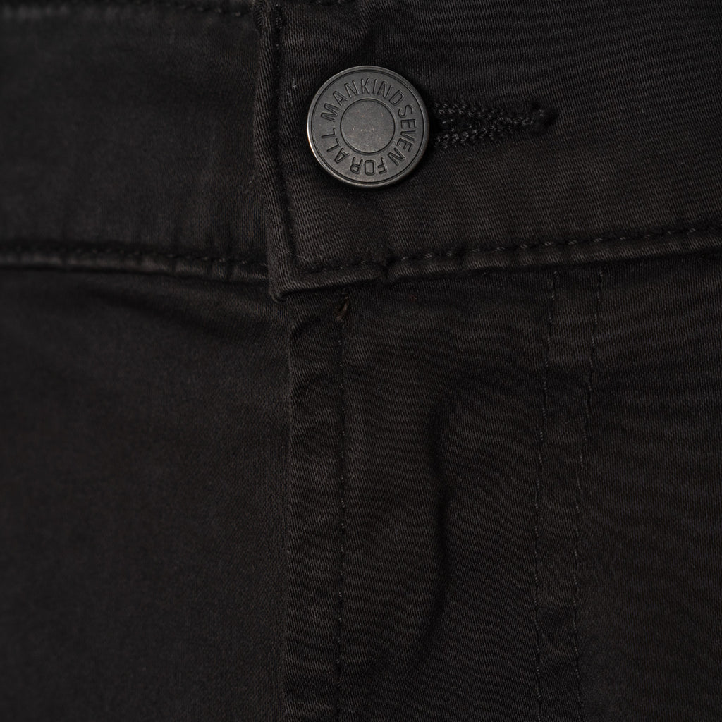 Close-up of black denim pants showing the button closure with a silver-toned button that reads '7 For All Mankind'.  7 For All Mankind Chinos available at Sakks Menswear in Southampton.