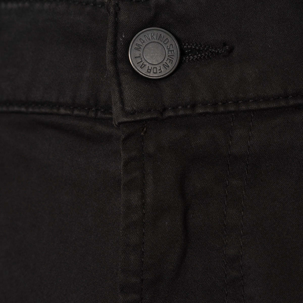 Close-up of black denim pants showing the button closure with a silver-toned button that reads '7 For All Mankind'.  7 For All Mankind Chinos available at Sakks Menswear in Southampton.