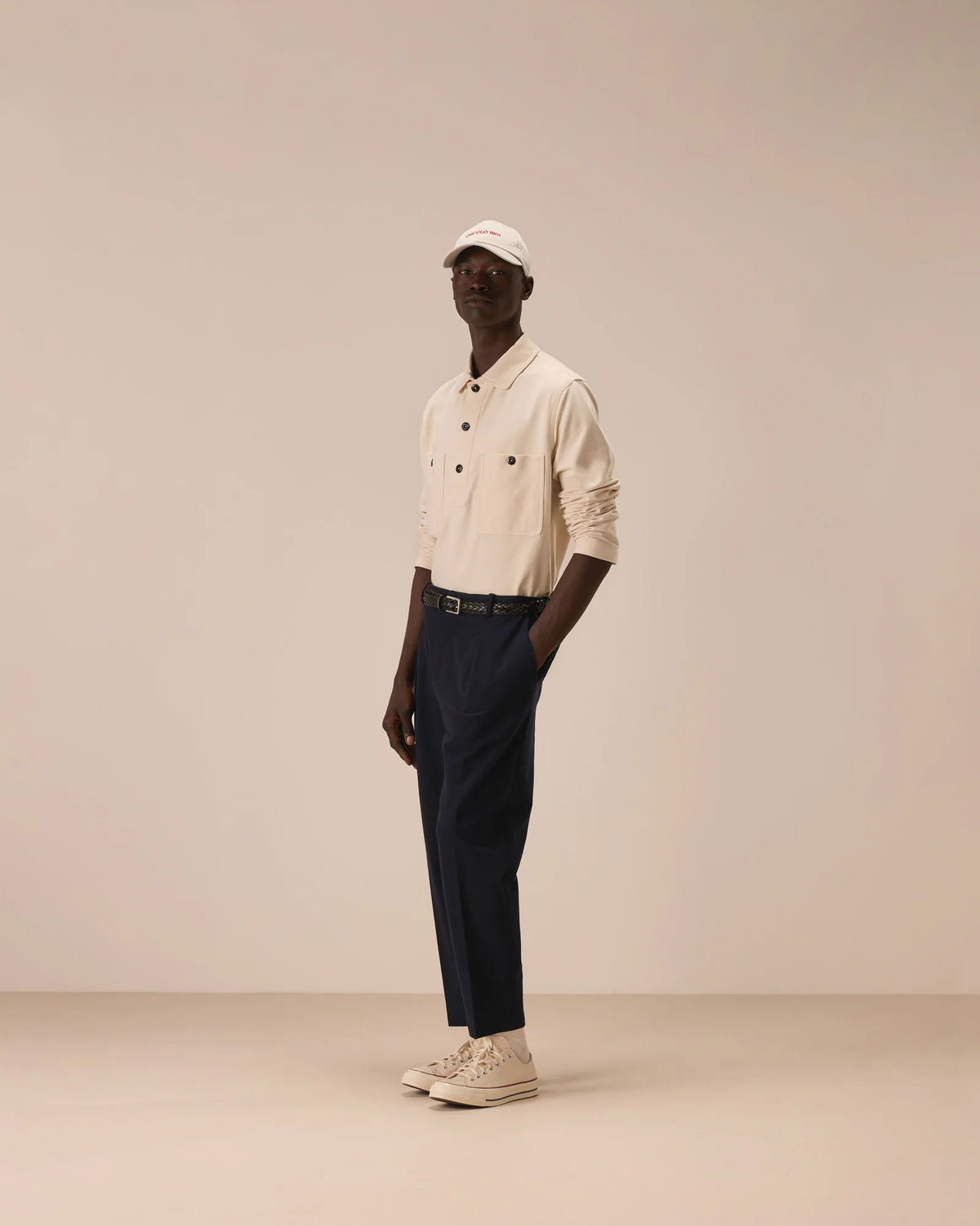 A model stands in profile wearing a cream-colored button-up shirt, dark navy trousers, a white cap, and off-white sneakers against a solid beige studio background. Circolo 1901 Sweatshirts available at Sakks Menswear in Southampton.