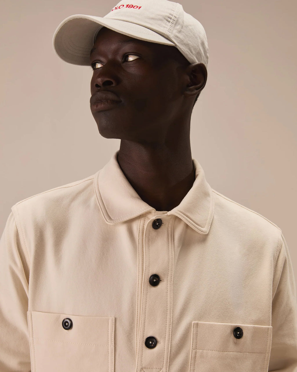 A man wearing a cream-colored collared button-up shirt and a matching cream baseball cap with red embroidered text, photographed against a solid beige background. Circolo 1901 Sweatshirts available at Sakks Menswear in Southampton.