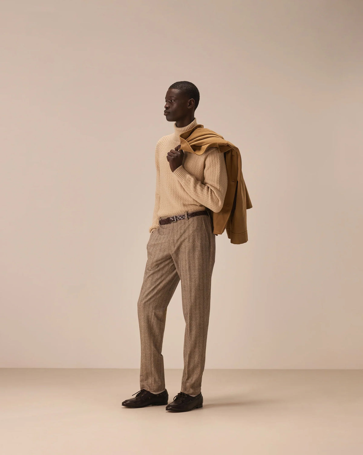 A man stands in profile wearing a cream knit sweater, beige plaid trousers, and black loafers, with a camel-colored coat draped over his shoulders, against a plain warm beige studio background. Circolo 1901 Trousers available at Sakks Menswear in Southampton.