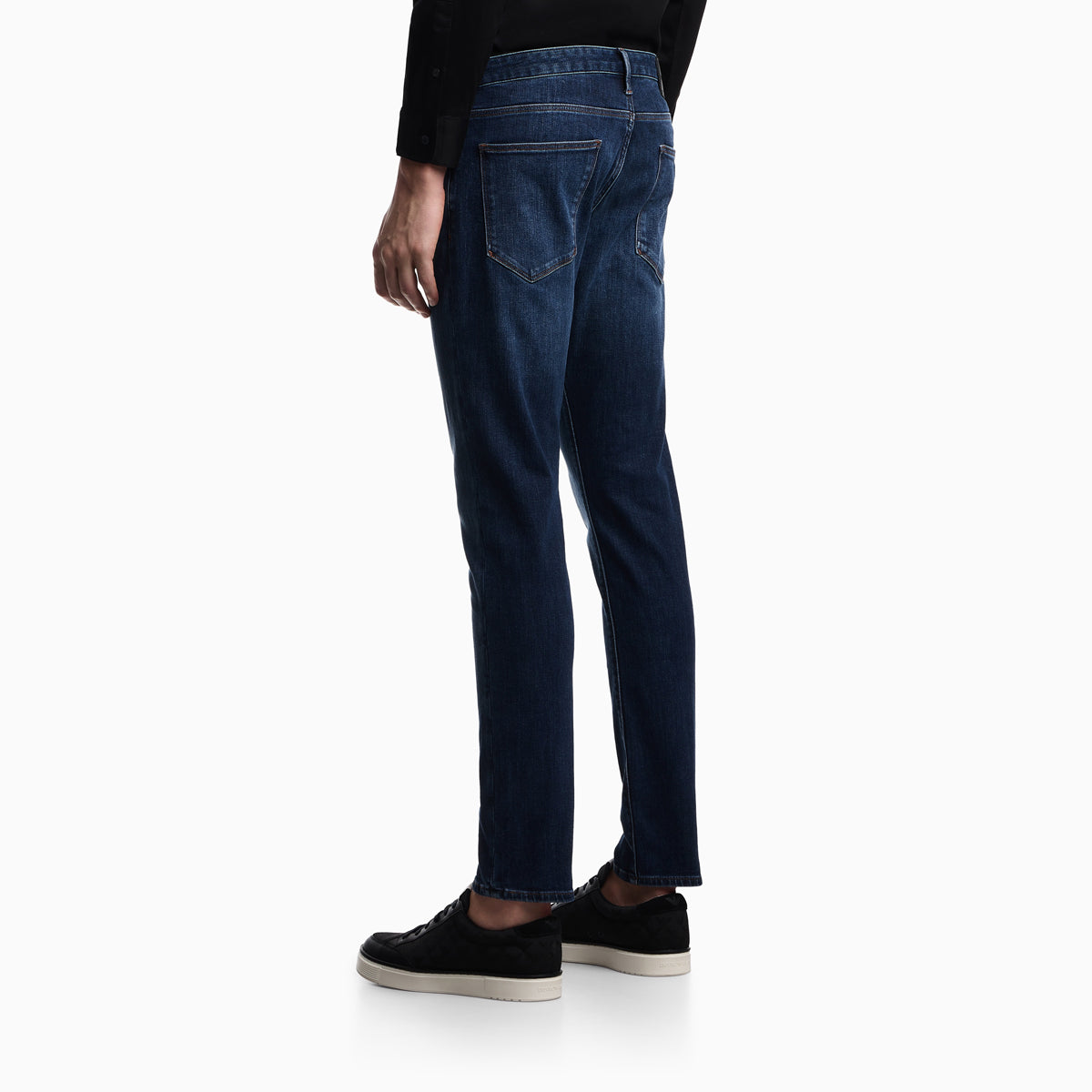A person shown from the back wearing dark blue slim-fit jeans, a black long-sleeve top, and black sneakers with white soles, against a plain white studio background. Emporio Armani Jeans available at Sakks Menswear in Southampton.