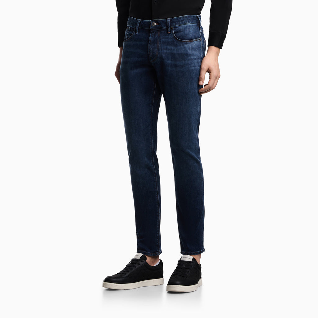 A person wearing dark blue slim-fit jeans with a black long-sleeve top and black low-top sneakers, shown from the waist down against a clean white studio background. Emporio Armani Jeans available at Sakks Menswear in Southampton.