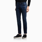 A person wearing dark blue slim-fit jeans with a black long-sleeve top and black low-top sneakers, shown from the waist down against a clean white studio background. Emporio Armani Jeans available at Sakks Menswear in Southampton.