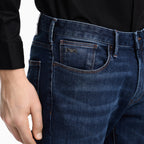 Close-up of a person wearing dark blue denim jeans with a black long-sleeve shirt. The jeans feature a visible front pocket with a small embroidered logo and contrast stitching. Emporio Armani Jeans available at Sakks Menswear in Southampton.